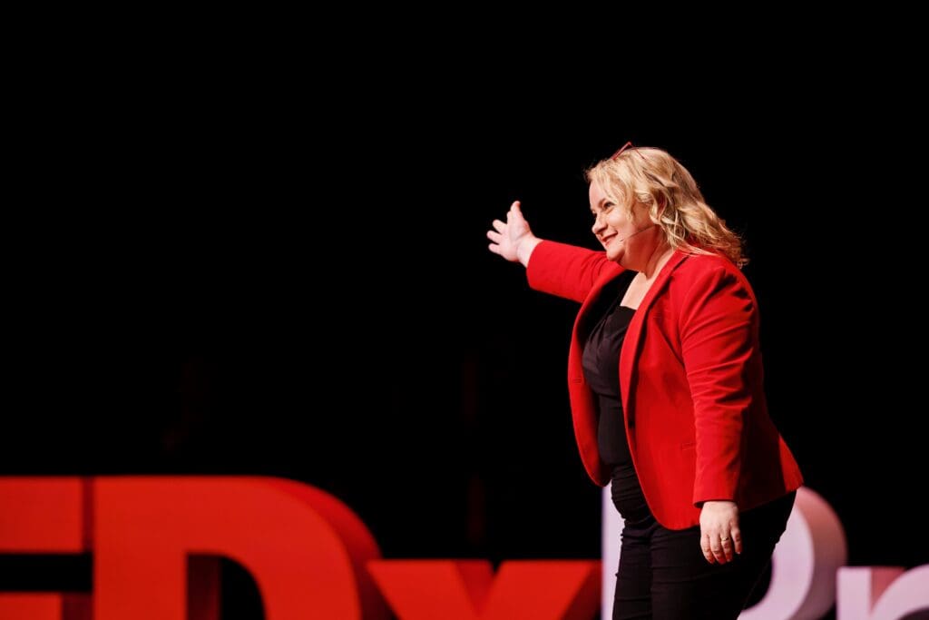 Juanita on stage as host of TEDxBrisbane, wearing a red jacket with her right arm raised towards side stage, inviting the audience to welcome the next speaker to stage with applause.