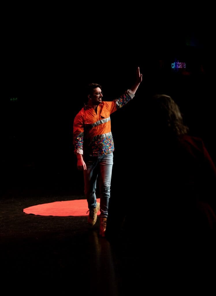 Daniel Allen, in his funky Trademutt shirt, walks off stage, smiling, with his left hand raised to acknowledge the standing ovation and applause of the audience.