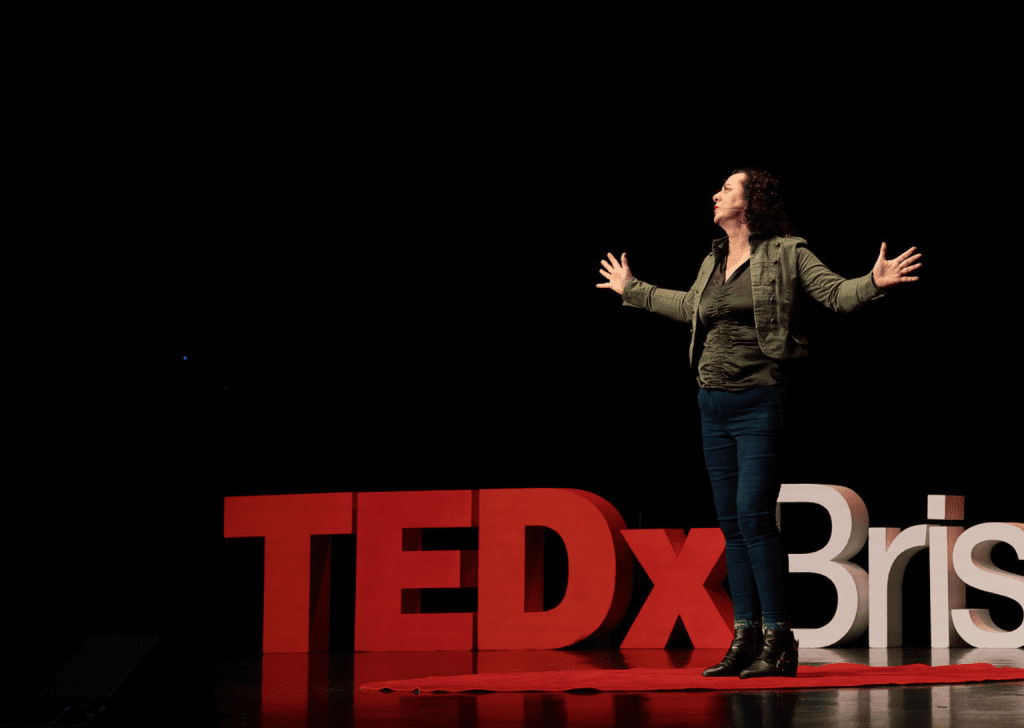 Adrienne Alexander wearing a green jacked and jeans delivering her TEDx Talk with both hands, open palms at 90 degrees to her side with palms facing front emphasising a point.